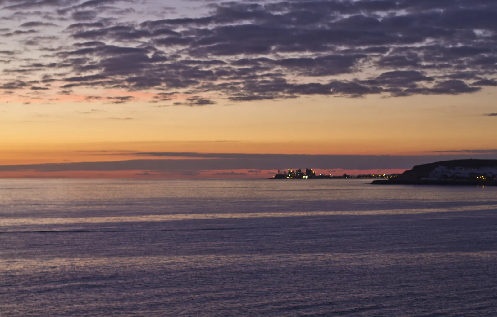 Calm sea at dusk off Arguineguín with an orange sky and the lights of the coast on the horizon