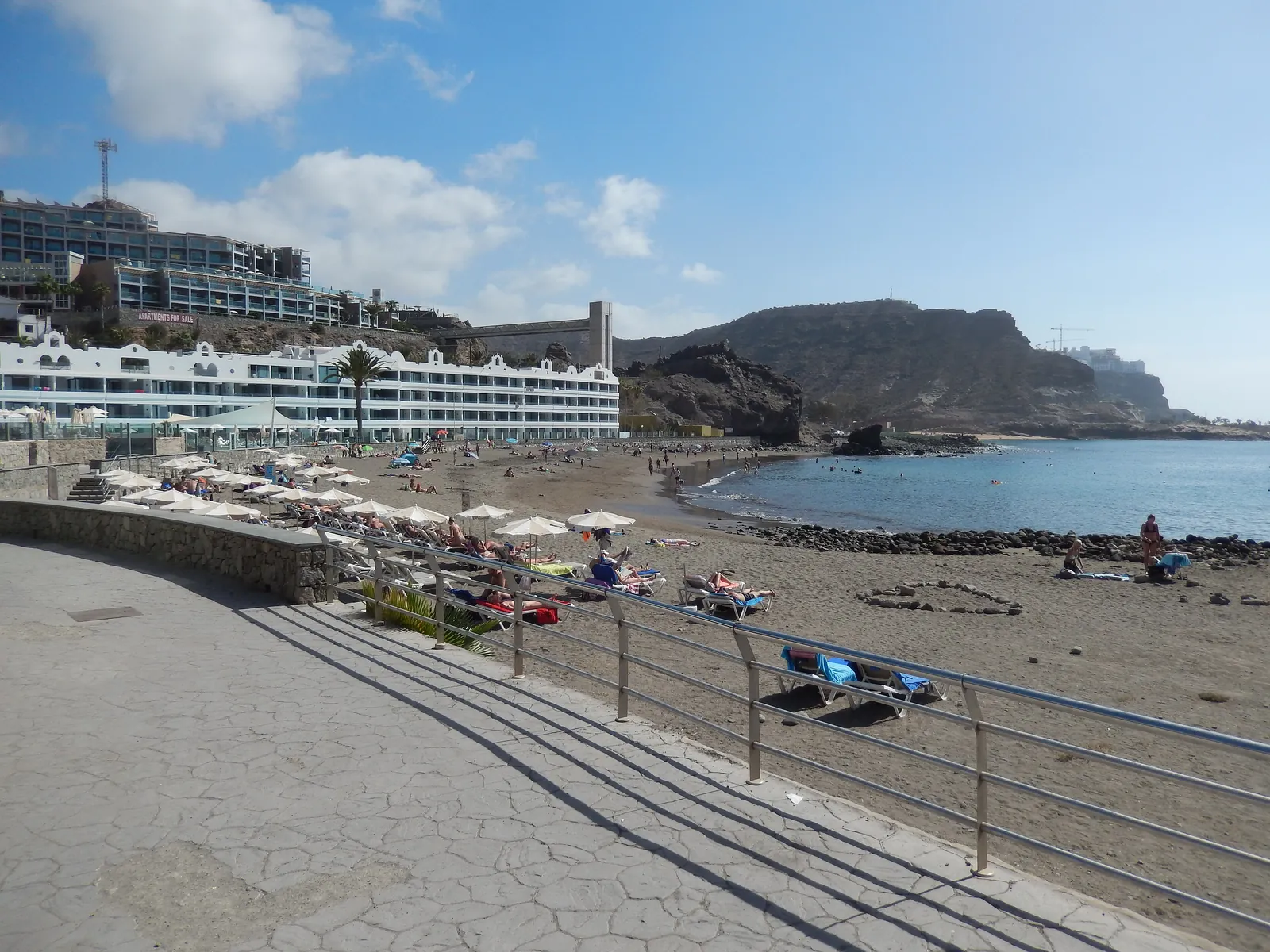 Dark-sand Playa del Cura seen from the railed promenade, with sunbathers and calm sea