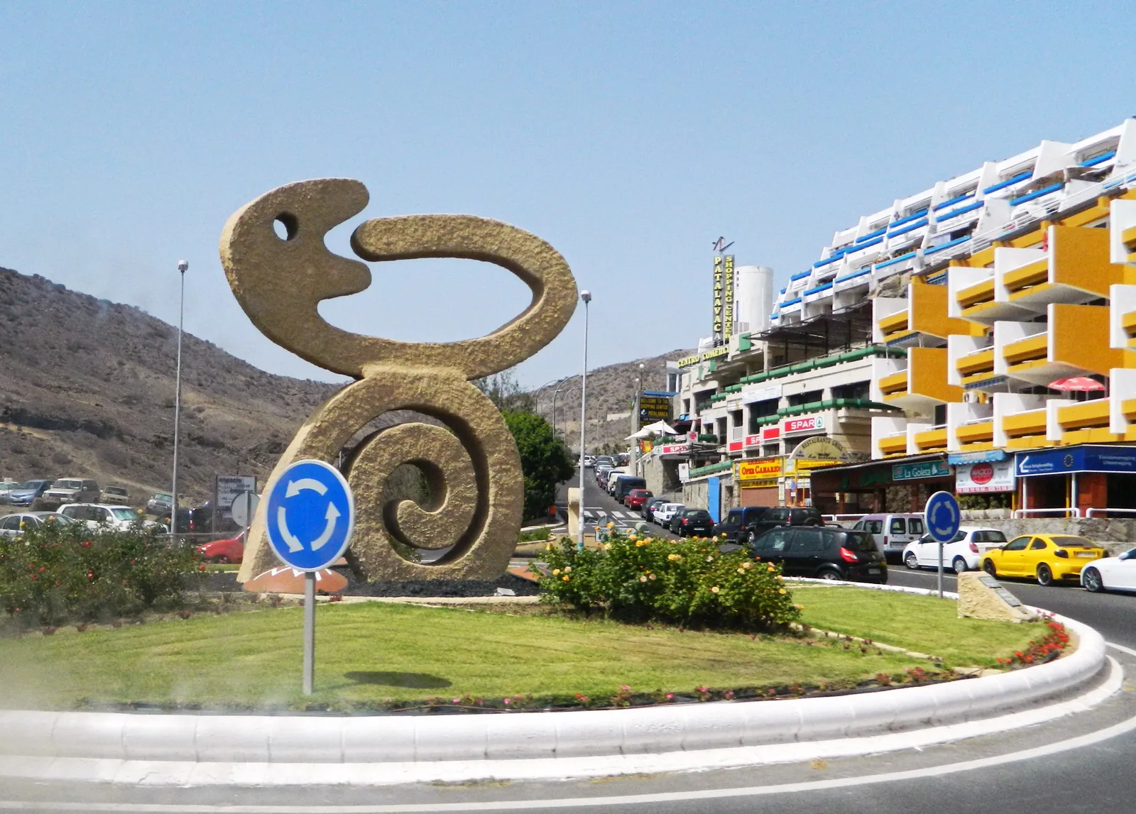 Sculpture on the roundabout at Patalavaca with the resort's shopping centre and apartment blocks beside the road