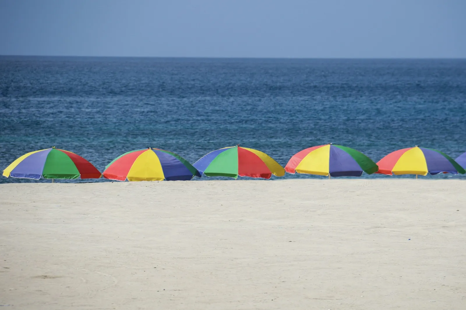 Row of colourful beach parasols on pale sand in front of a calm deep blue sea