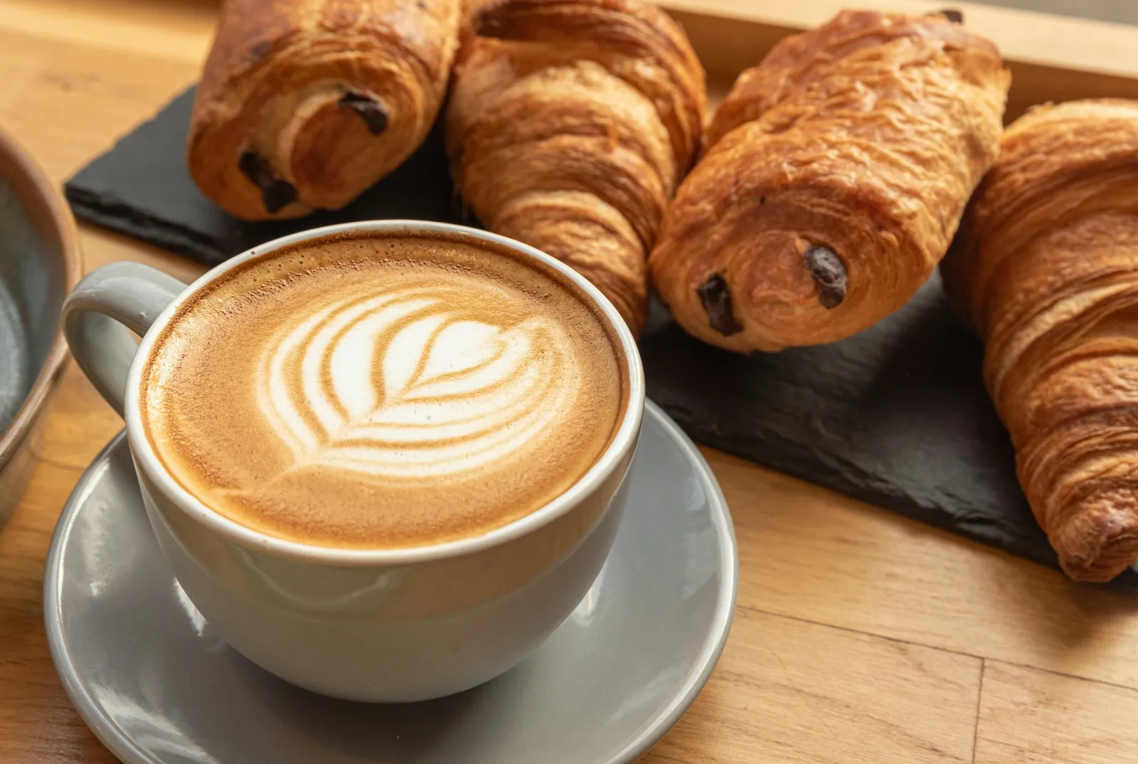 Latte with leaf art in a grey cup beside fresh croissants and pain au chocolat on a slate board
