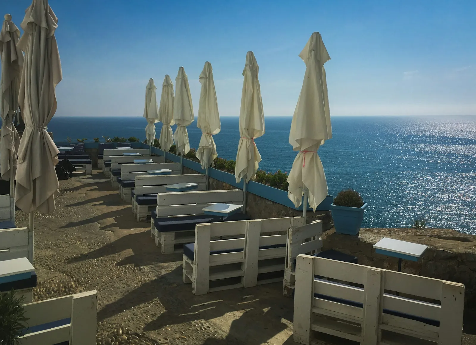Terrace with closed parasols and white benches above a sparkling blue sea