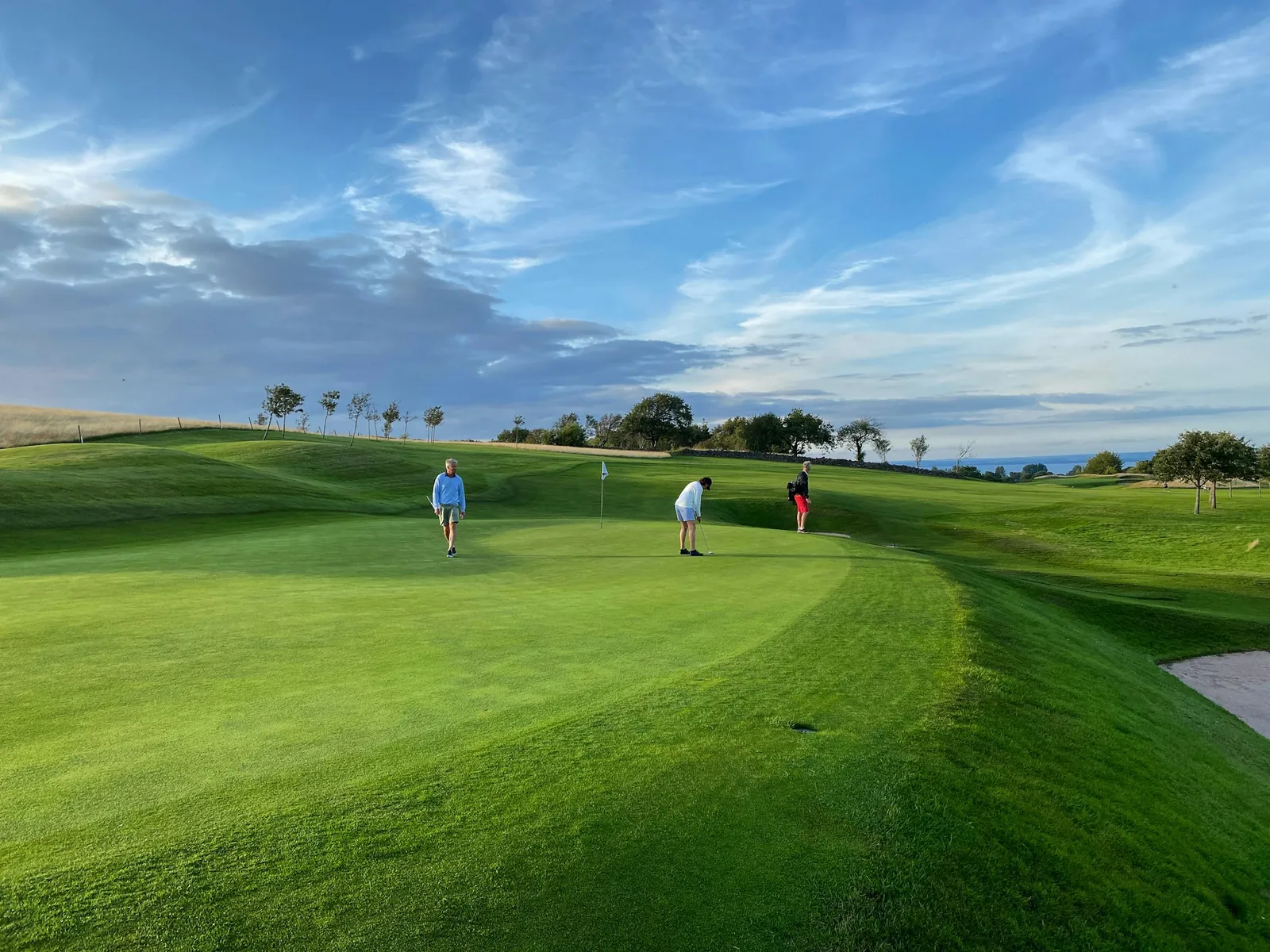 Golfers putting on a green fairway under a blue sky with sea on the horizon