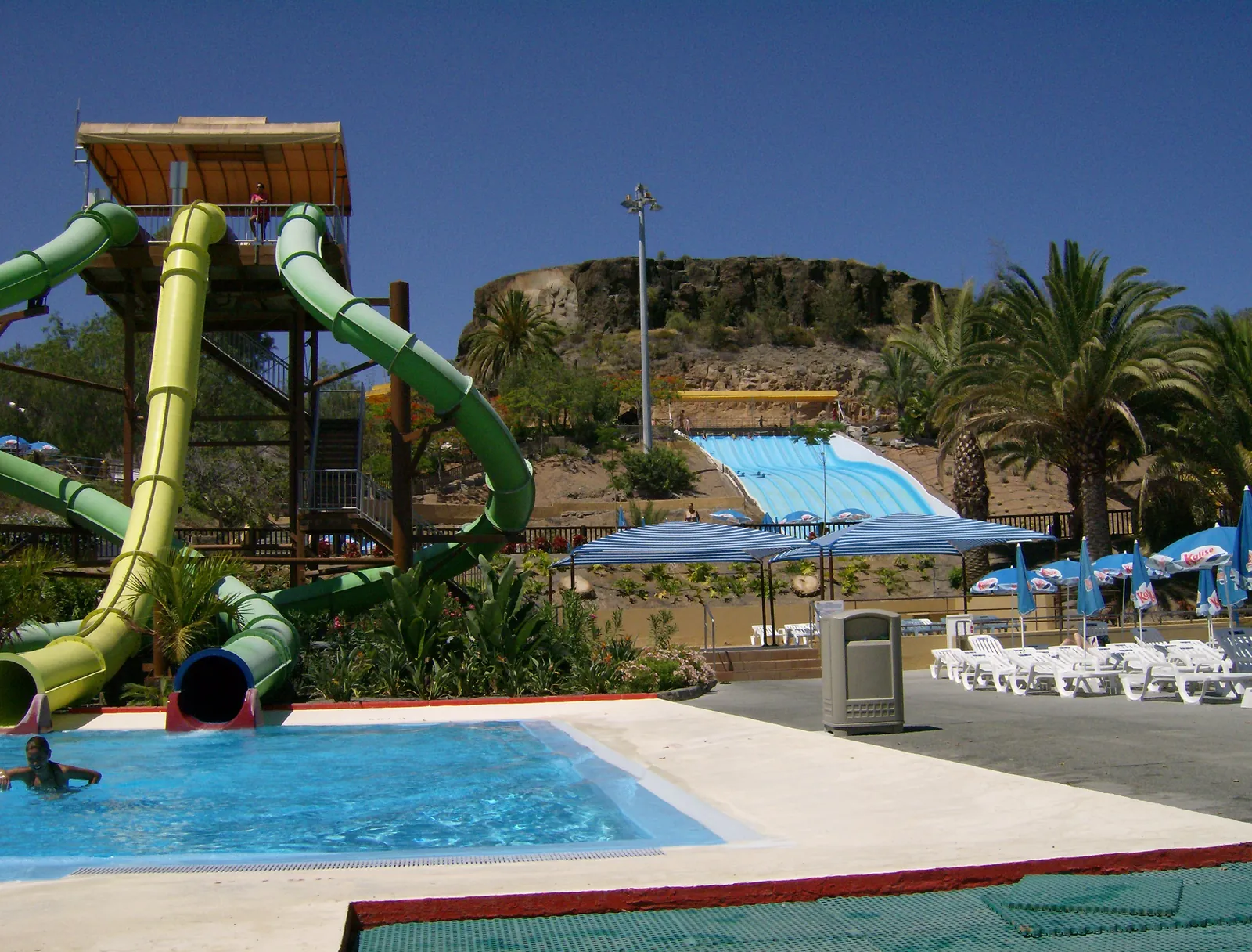 Green and yellow water slides above a pool at Aqualand Maspalomas with sun loungers and palms behind