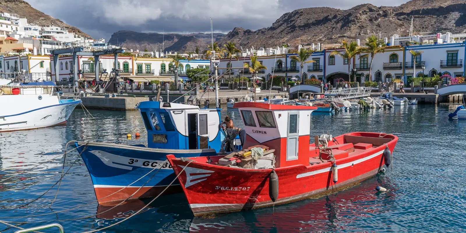 Red and blue fishing boats moored in Puerto de Mogán harbour below white houses and dry mountains